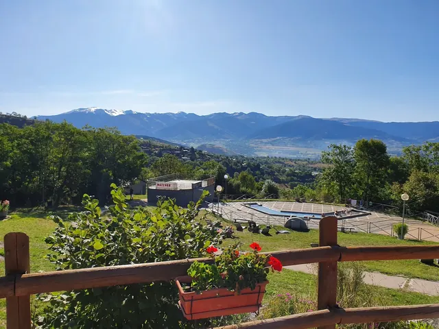 Wide view of the Bains de Dorres hot spring pool on a sunny day with green trees, grass, flowers, and distant mountains near Font-Romeu, Fr.
