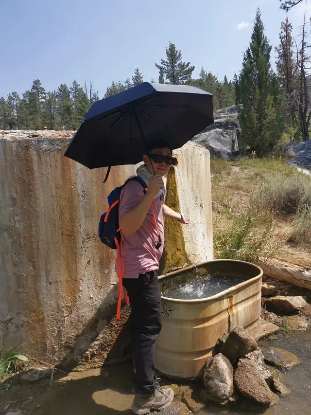 Man holding a black umbrella stands beside a rust-streaked basin with flowing water at Mono Hot Springs in the forested area of California.