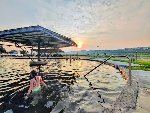 People bathing in the outdoor pool at Riverdale Resort Hot Springs at sunset near Preston, Idaho, with a metal railing and open landscape.
