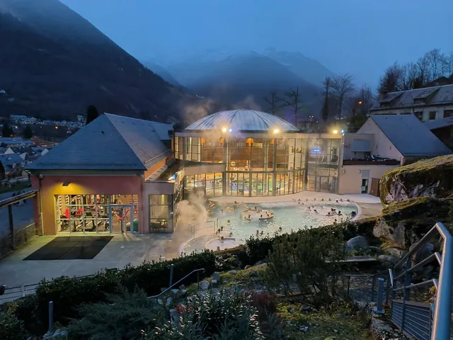 Evening view of the outdoor and indoor hot spring pools at Balnea in Loudenvielle, France with steam, surrounding buildings, and mountains.