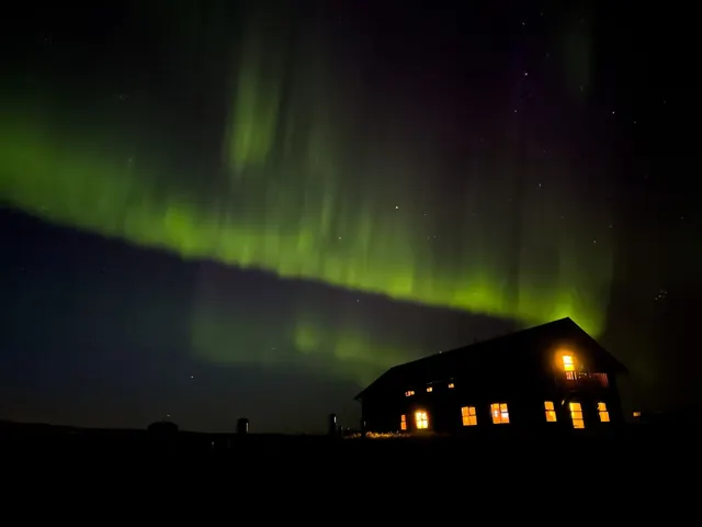 Dark silhouette of a building lit from inside with northern lights glowing bright green and purple in the night sky above in Iceland.