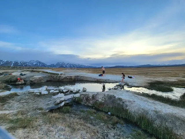 Visitors walking alongside connected soaking pools bordered by rocks and a wooden boardwalk at Crowley Wild Willy's Hot Spring with a wide.