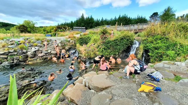 People soaking in a natural rock pool hot spring near a small waterfall with lush greenery and a wooden fence in Taupō new-zealand.