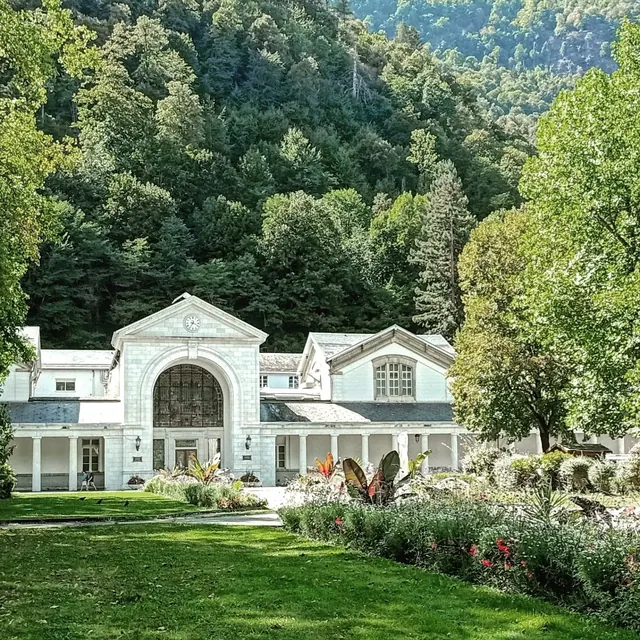 Historic white stone building with arch entrance surrounded by lush green trees and flowering garden at Thermes de Luchon, France.