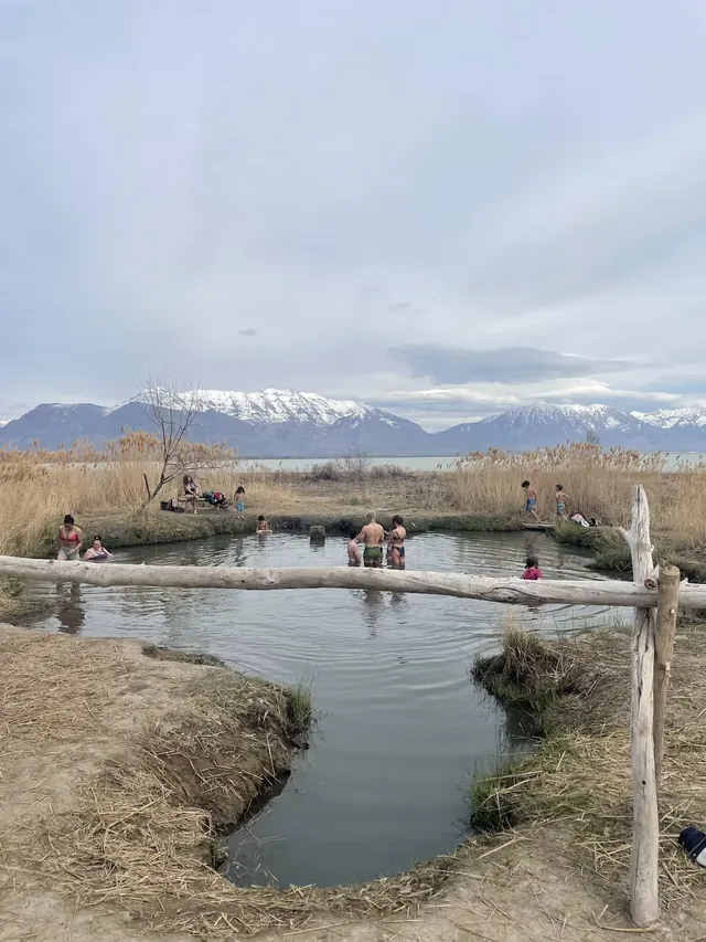 Visitors soak in a calm hot spring pool surrounded by grasses and a wooden fence with snowy mountains visible in the background near Sarato.