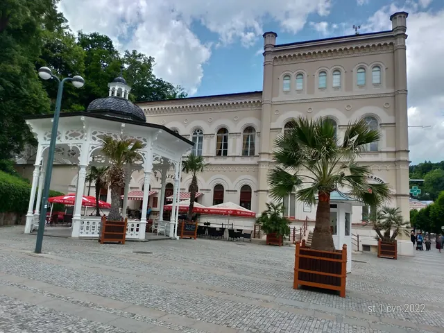 White ornate pavilion with palm trees in large stone-paved square outside Windsor SPA Hotel in Karlovy Vary Czechia.