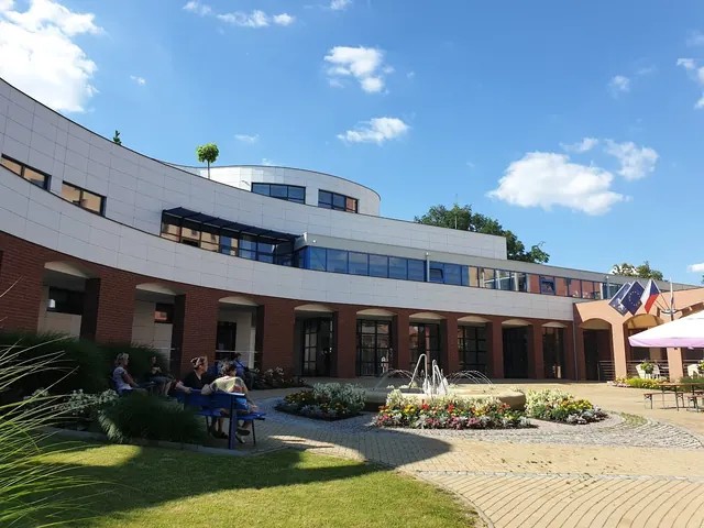 People relaxing on benches beside a central fountain in Lazne Hodonin outdoor courtyard under a partly cloudy sky in Hodonin, Czechia.