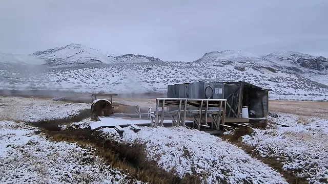 Snow-covered Alvord Hot Springs bathhouse and pool with visible steam, surrounded by snowy hills and white winter landscape near Princeton.