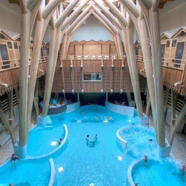 Large indoor mineral pool with people relaxing in water under wooden arches inside Aquensis Spa Thermal in Bagneres-de-Bigorre France.
