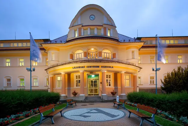 The exterior of Lazeanske centrum Agricola building at dusk with warm lights, pink columns, benches, and two flagpoles in Jachymov Czechia.