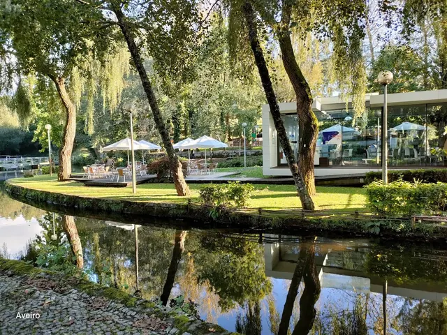Green park area with white parasols and a glass cafe building beside a reflective water canal at Termas de S. Jorge by Santa Maria da Feira.