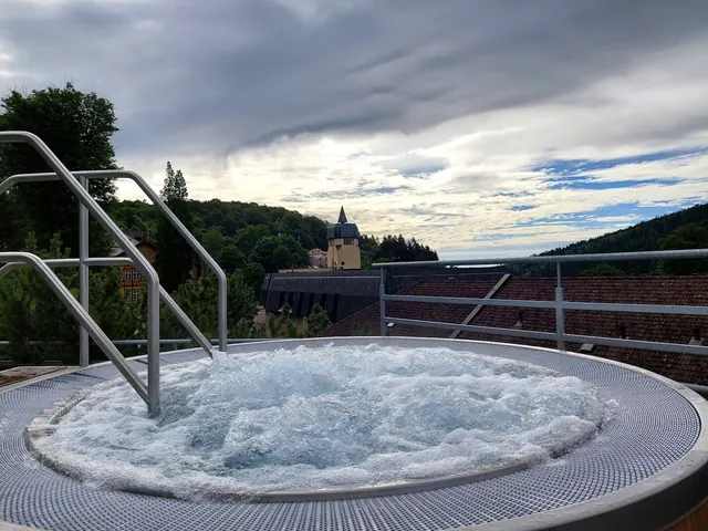 Outdoor bubbling hot tub with metal railing overlooking rooftops and a tower in Janske Lazne, czechia under cloudy sky.