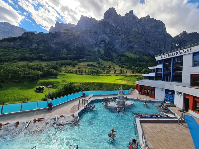 Crowded outdoor spa pool at Leukerbad Therme with people relaxing near a modern fountain under a partly cloudy sky and steep green mountain.