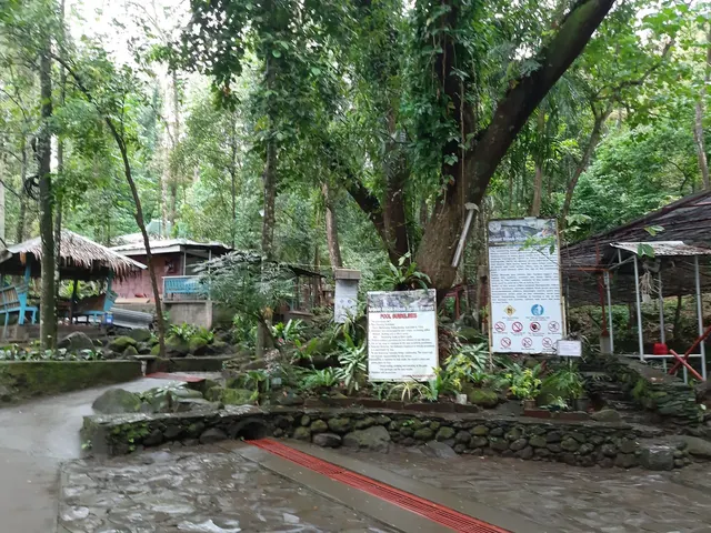 Pathway and signs near shaded seating areas and huts surrounded by trees at Ardent Hot Spring in Mambajao.
