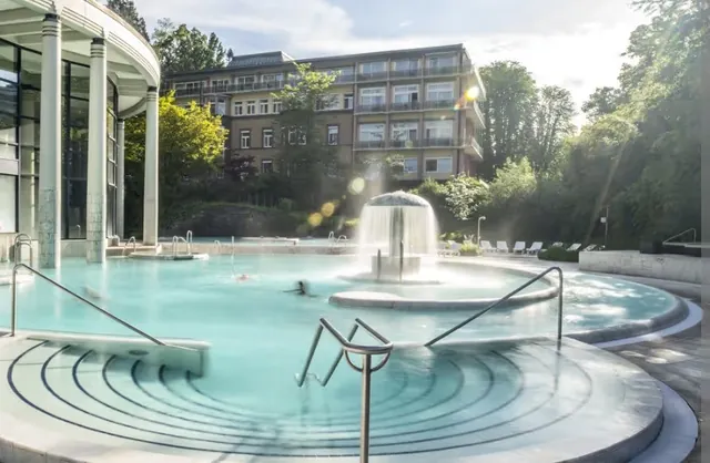 Sunlit outdoor mineral pool at Caracalla Therme with a central water feature and curved steps leading into the water.