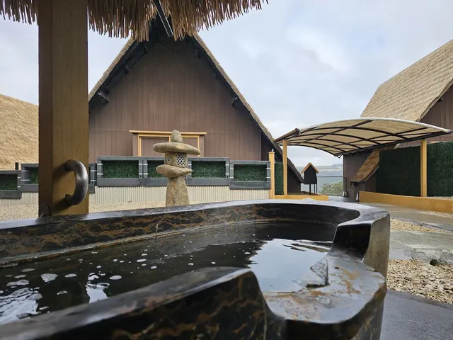 Outdoor hot spring pool with stone lantern sculpture beside traditional thatched roof buildings under a blue sky in South Bowenfels Austral.