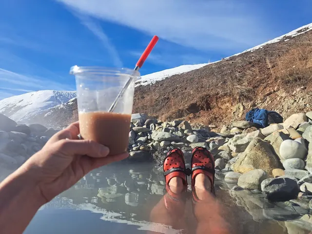 Steaming natural hot spring soaking pool surrounded by rocks with a hand holding a drink and feet wearing red shoes at Astoria Hot Springs.