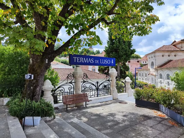 Sign reading Termas de Luso mounted on decorative black fencing near steps under a large leafy tree in Luso, Portugal.