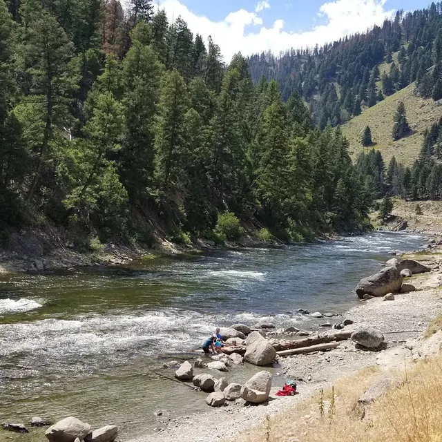 A natural hot spring area beside a flowing river with people enjoying the water and pine trees on hills at Sunbeam Hot Springs, Idaho.