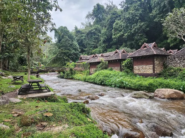 River flowing through the hot spring resort area with rustic buildings and dense forest in the background at Chae Son Hot Springs, Thailand.