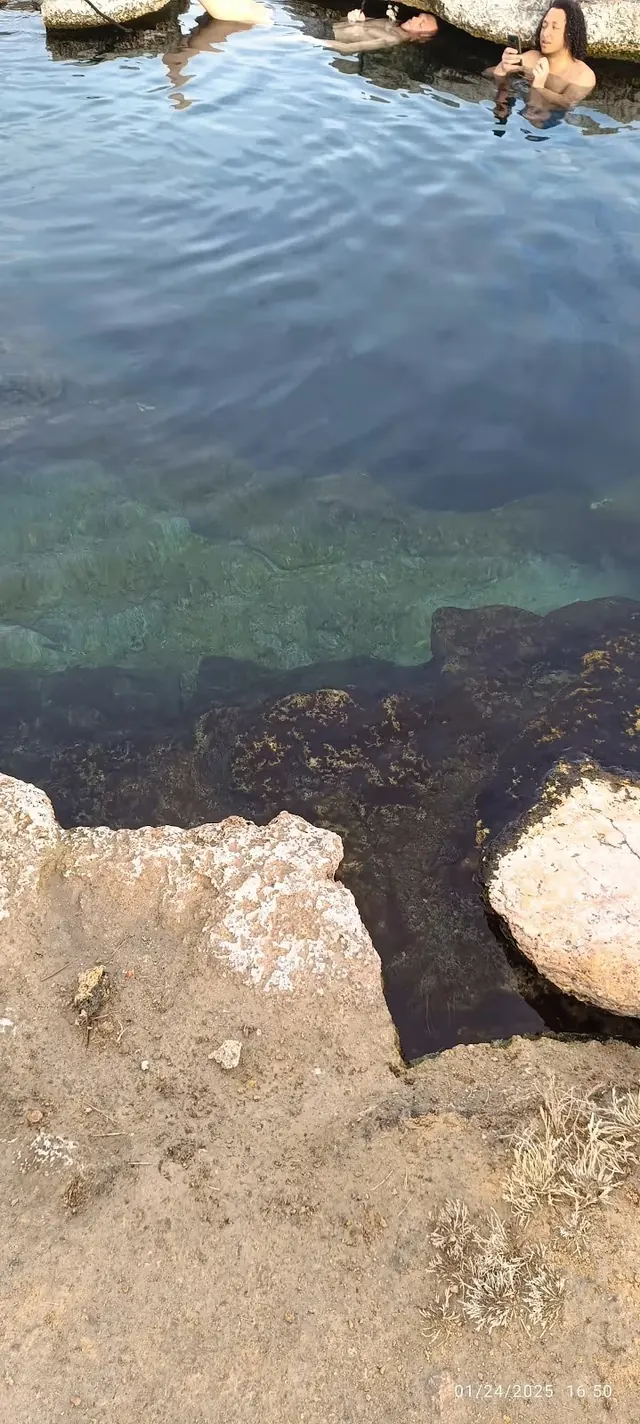 Soaking pool at Meadow Hot Springs with individuals relaxing near rocky ledges and clear mineral water visible below.