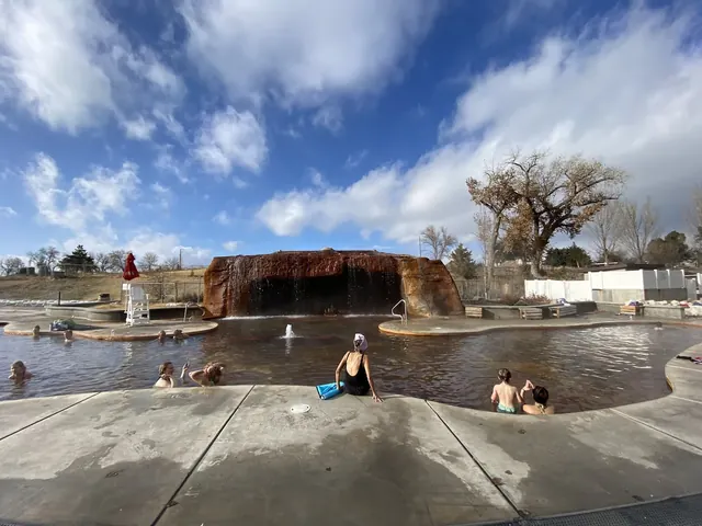 Outdoor Roosevelt Hot Springs pool with a rock waterfall feature and visitors soaking under a partly cloudy sky near Milford Utah.