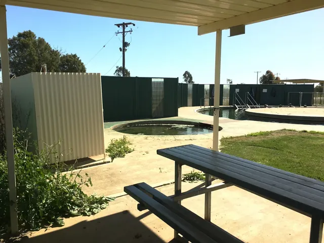 Shaded picnic table with outdoor seating next to multiple hot spring pools enclosed by green fencing at Burren Junction Artesian Bore Baths.