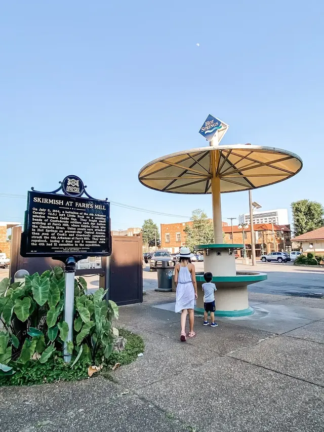 A woman and child walking toward the Hill Wheatley Plaza Jug Fountain under clear blue skies in the urban center of Hot Springs, Arkansas.
