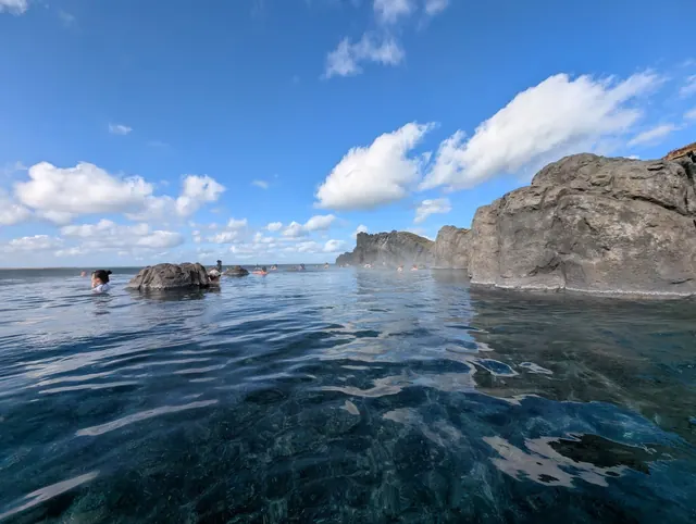 Visitors soak in the natural outdoor hot spring surrounded by large rocks with steam rising under a clear blue sky at Sky Lagoon near Reykj.
