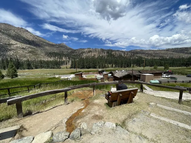 Overlooking Grover Hot Springs pool area from a hilltop bench with a flowing mineral stream and mountain ridges under a partly cloudy sky.