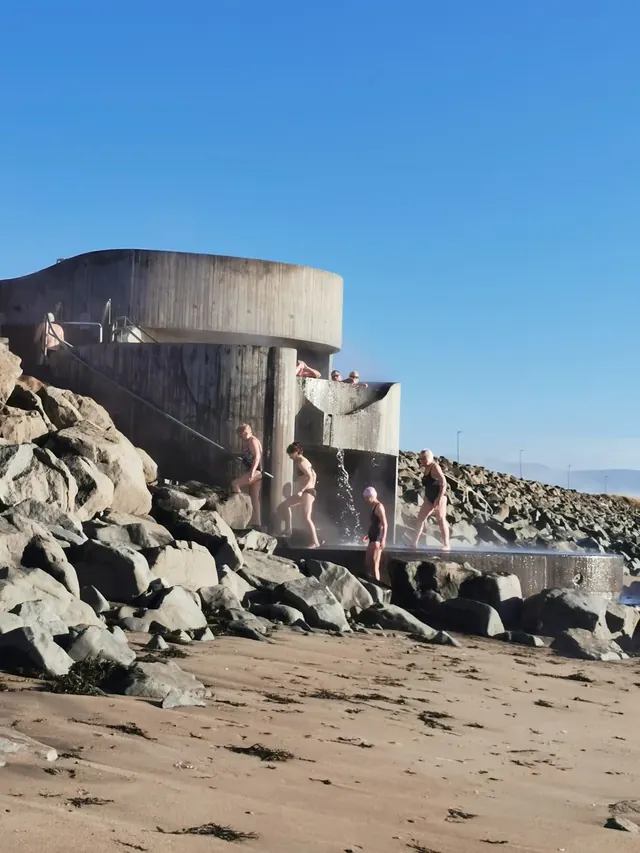 Group of people enjoying a hot spring bath next to a spiral concrete structure at Gudlaug Baths in Iceland.