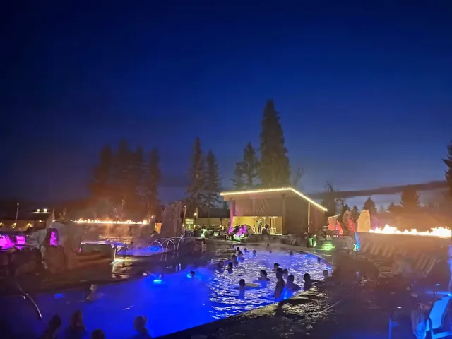 Night view of Bozeman Hot Springs outdoor pool with people soaking, colorful lighting, and fire features in Montana.