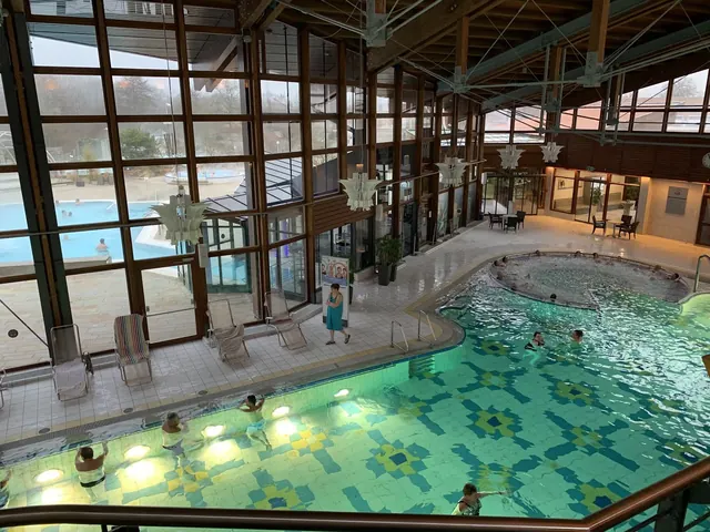 Indoor pool area with patterned tiles and several people bathing under a wooden ceiling with large windows at Obermain Therme in Germany.
