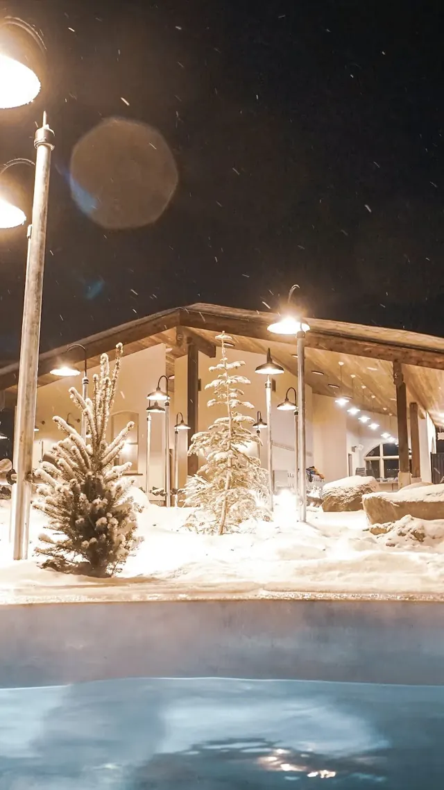 Nighttime view of The Springs hot spring pool with steam rising under glowing lamps and snowy small pine trees.