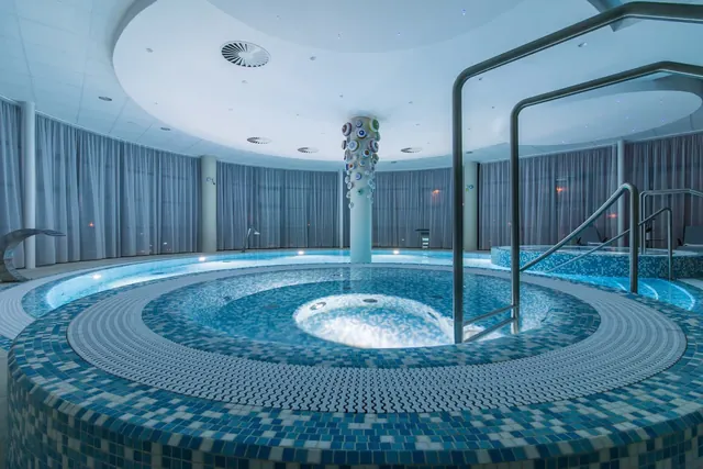 Indoor circular hot spring pool at Lazne Hodonin spa in Hodonin, Czechia, featuring mosaic tiles and metal handrails.