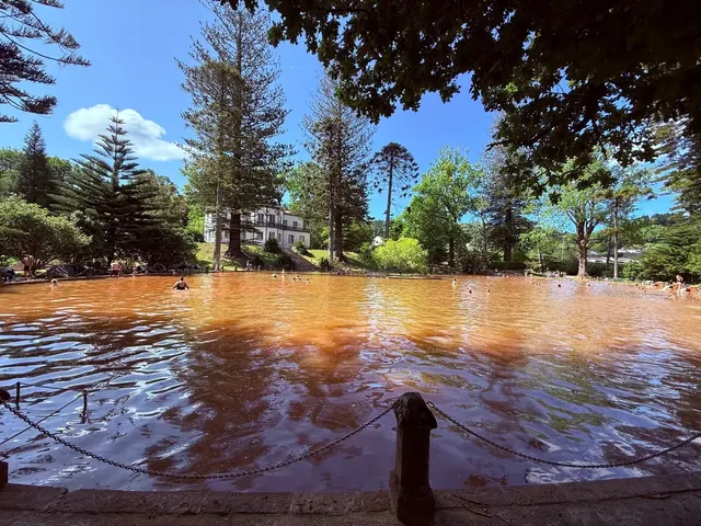 Terra Nostra Thermal Pool shows a rich orange soaking pool near a white building surrounded by pine trees in Furnas, Portugal.