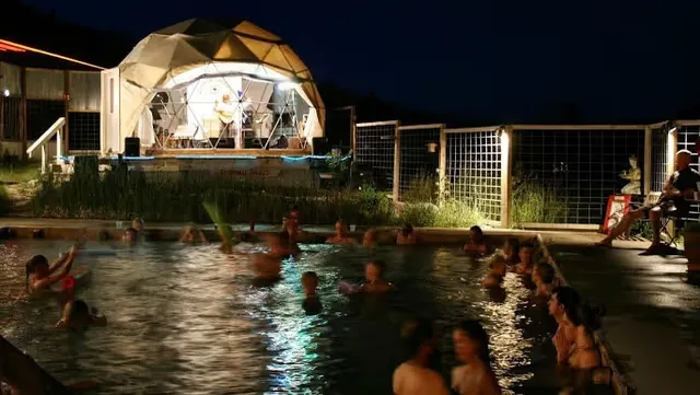 Night view of Norris Hot Springs with lit domed structure beside a crowded outdoor hot spring soaking pool in Norris Montana.