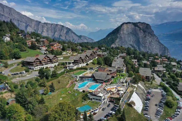 Aerial view of Les Bains d'Ovronnaz hot spring complex with pools near alpine village and forested mountain slopes in Switzerland.