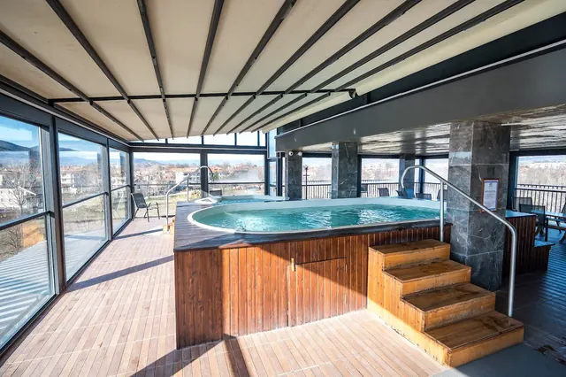 Indoor spa hot tub with wooden paneling and steps, located in a bright room with large windows overlooking the city of Banya, Bulgaria.