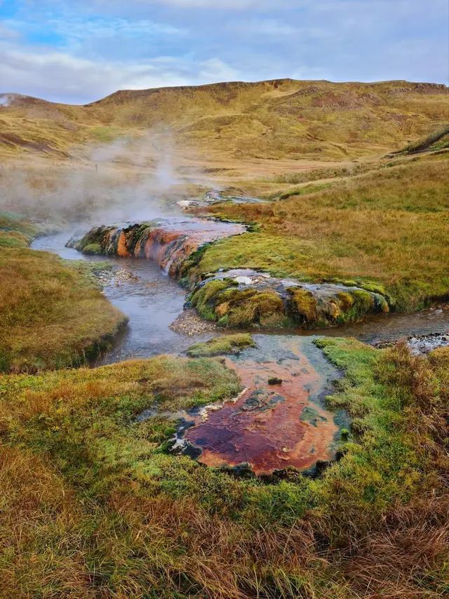 Steam rising from a colorful mineral-rich hot spring flowing through mossy grassland with hills in the background near Hverageroi, Iceland.
