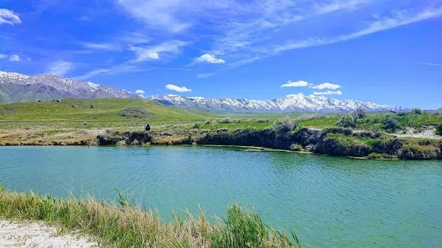 Large mineral pool with greenish water reflecting blue sky and snow-capped mountains visible beyond the shore in Horseshoe Springs Utah.