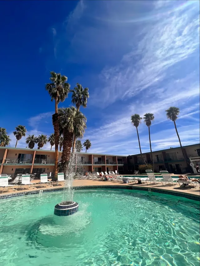 Fountain sprays water in the center of a turquoise-tiled outdoor spa pool with sunlit palm trees and hotel balconies in Desert Hot Springs.