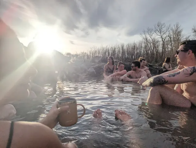 Group of people enjoying a steaming hot spring pool with the low sun and leafless trees in the background at Renova Hot Springs, Montana.