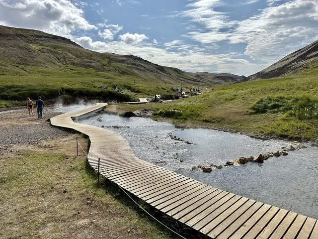 A winding wooden boardwalk beside a steaming hot spring river with visitors walking and soaking near Hverageroi in Iceland.