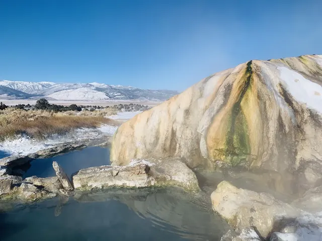 Natural hot spring pool with steaming water and colorful travertine mineral formation near Bridgeport, California under clear blue sky.