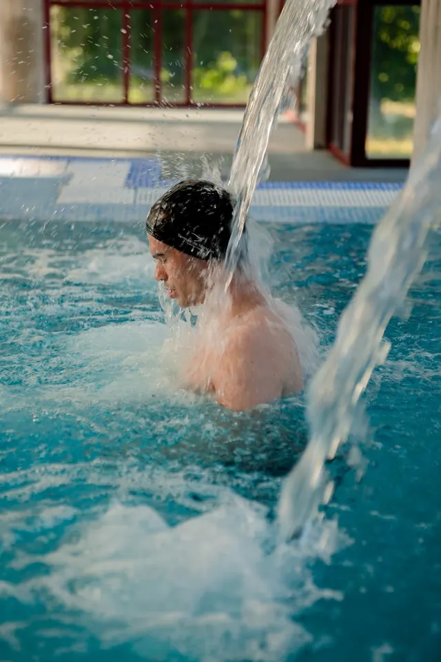 Close-up of a man wearing a swim cap standing under cascading water jets inside the indoor pool at Termas de Moncao near Moncao Portugal.