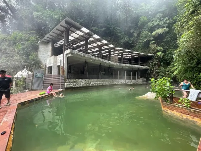 Outdoor hot spring with cloudy mineral water in a large pool beside a covered pavilion surrounded by misty rainforest at Las Fuentes Georgi.