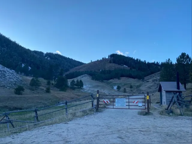 Entrance gate with informational signs to Worswick Hot Springs, set against forested hills under a clear blue sky near Fairfield, Idaho.