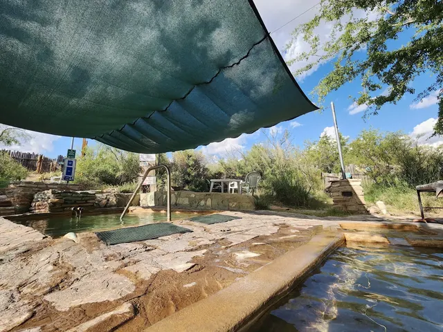 Two heated mineral pools at Faywood Hot Springs under a large shade cloth with a stone walkway and outdoor seating in New Mexico.