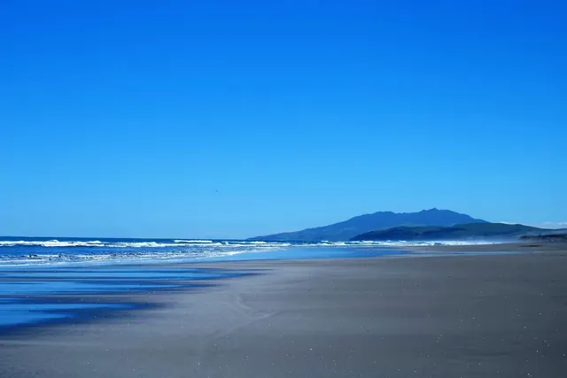 Wide sandy beach with gentle waves and distant mountain range under a vivid blue sky near Kawhia Hot Springs in Kawhia New Zealand.